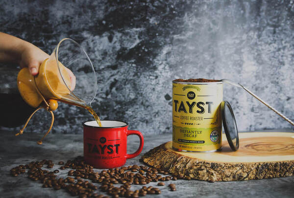 Coffee being poured into a Tayst mug alongside a tin of Tayst coffee.