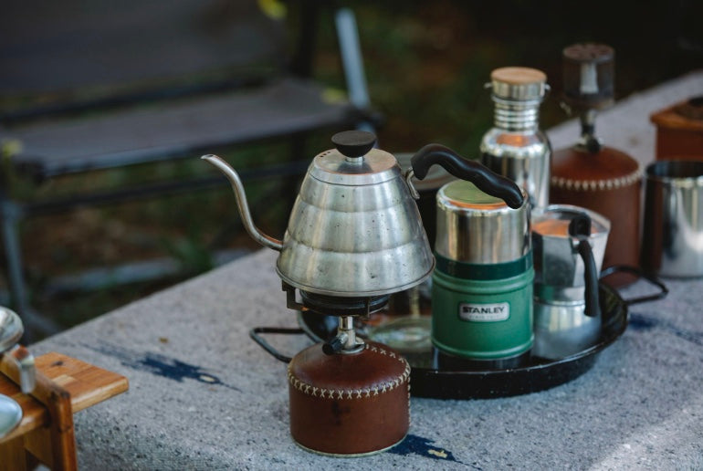 A camp coffee-making setup including a kettle, a portable stove, and a thermos. 