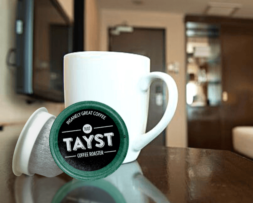 White ceramic mug with a Tayst coffee capsule in front, placed on a reflective table with a softly blurred indoor background.