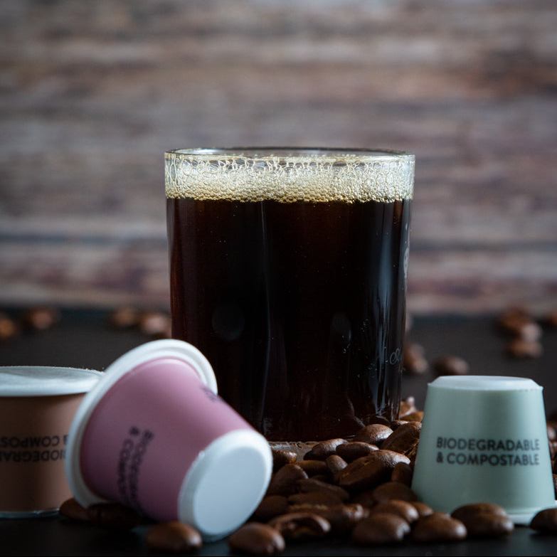 Glass of dark liquid with foam, surrounded by coffee beans and tayst biodegradable nespresso cups on a wooden surface.