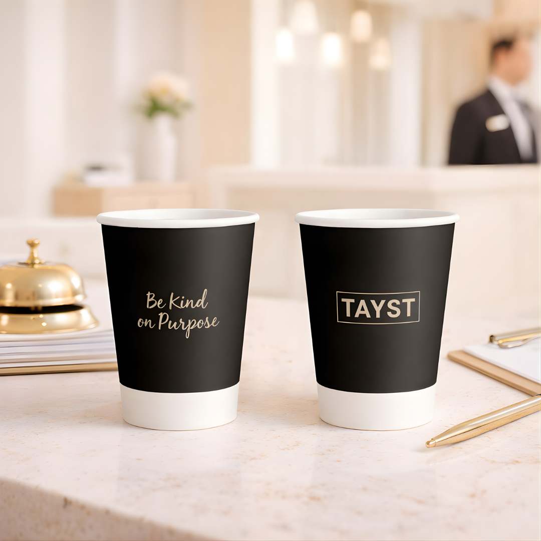 Two black coffee cups with 'Be Kind on Purpose' and 'TAYST' text on a blurred background.