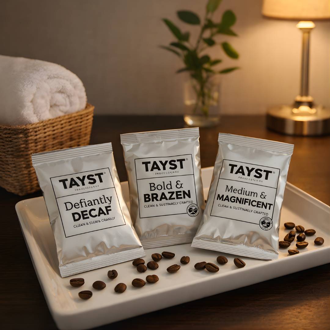 Three TAYST coffee frac packs on a tray with scattered coffee beans, in a hotel setting.