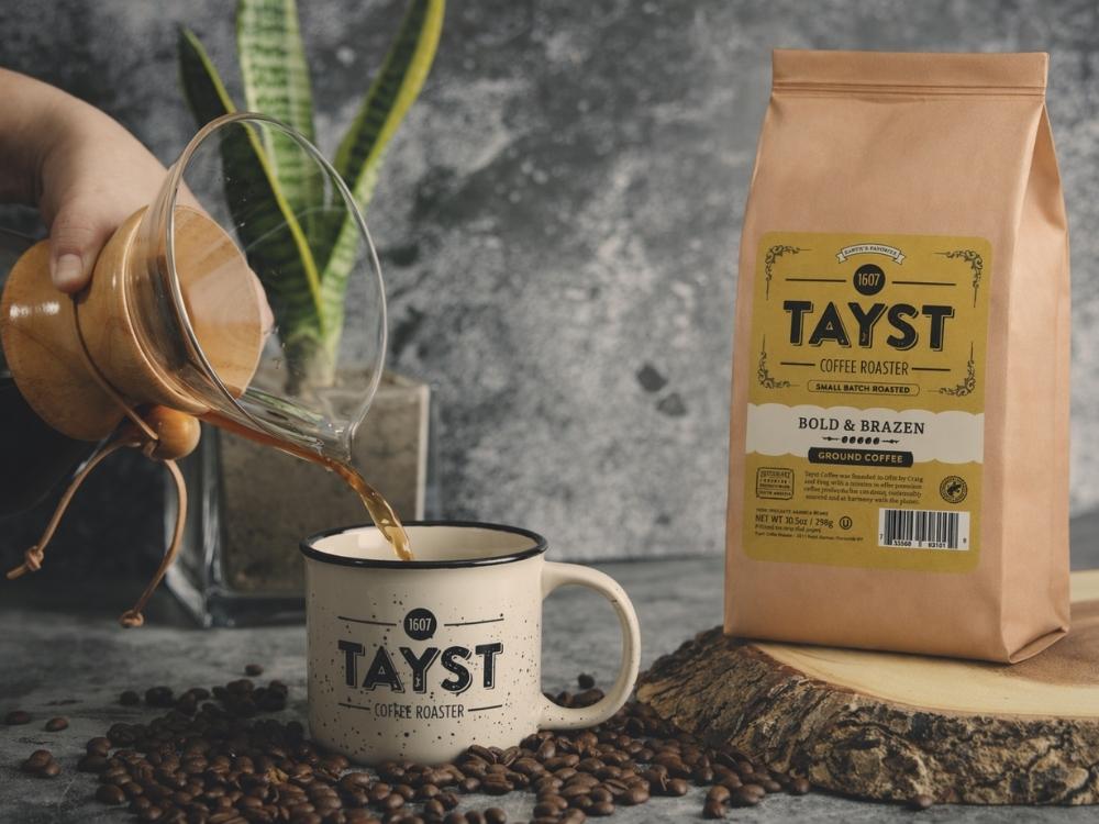 Person pouring coffee from a glass pot into a white mug with 'TAYST' branding, next to a bag of TAYST ground coffee beans.