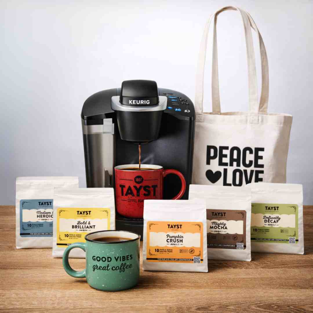 Keurig coffee maker brewing Tayst coffee with assorted coffee pods, two branded mugs, and a tote bag on a wooden surface.